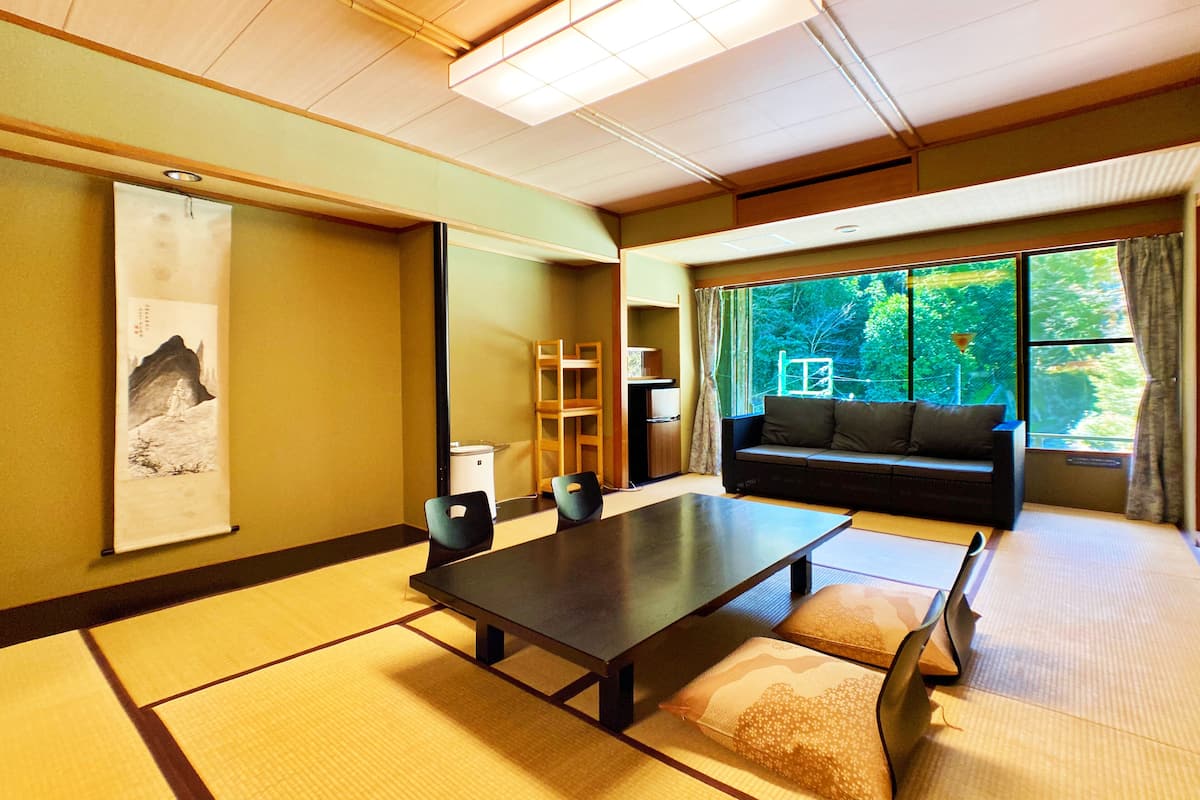 Habitació japonesa tradicional amb sòl de tatami, una taula baixa, un sofà i una finestra gran amb vista a vegetació.