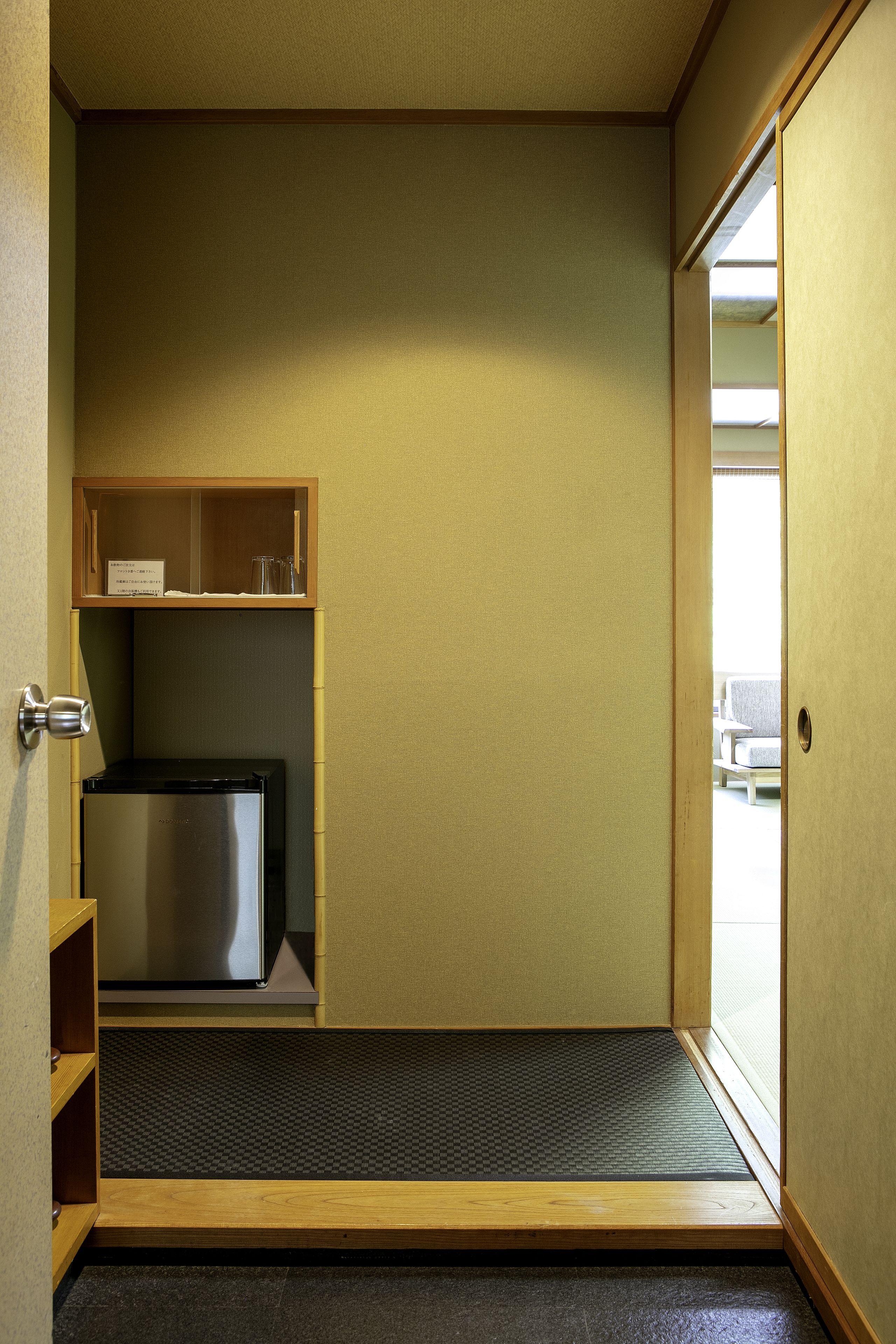 Habitación compacta con puerta corredera, un pequeño armario de madera, un refrigerador y un felpudo en la entrada.