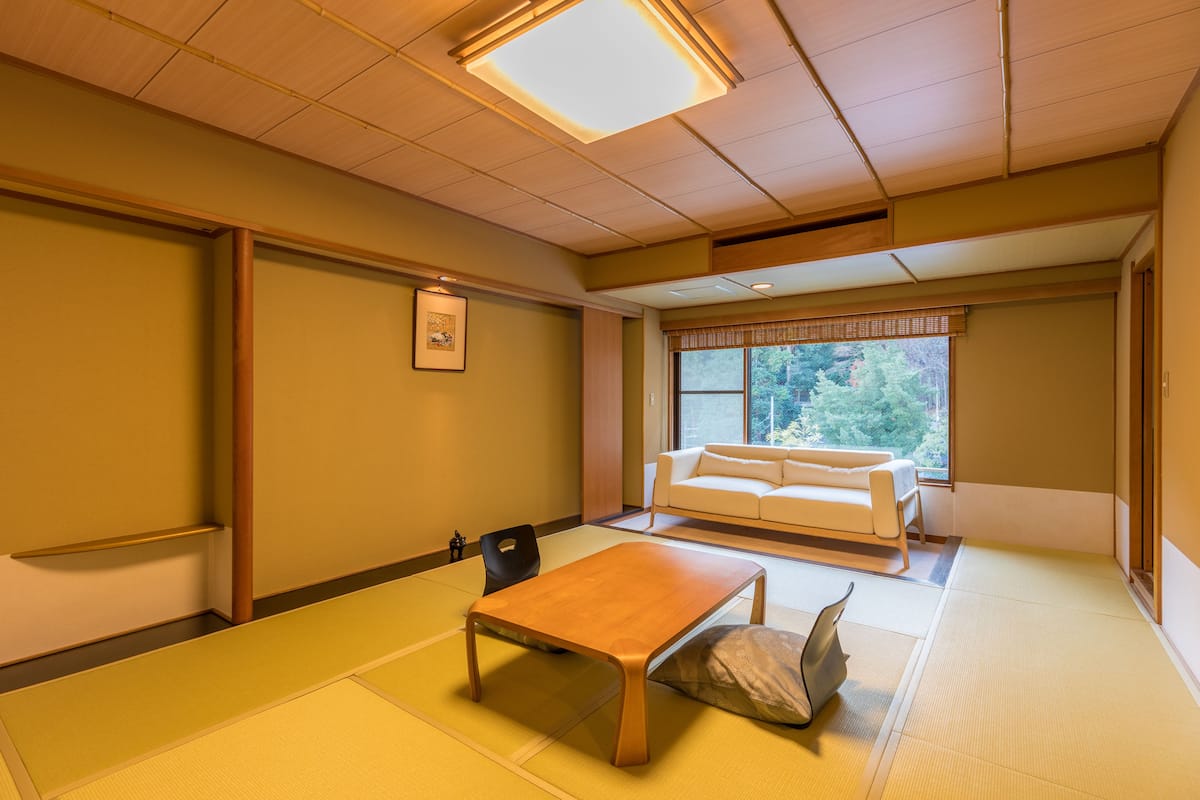 Habitació japonesa tradicional amb sòl de tatami, taula de fusta baixa, sofà i finestra amb vista a vegetació.