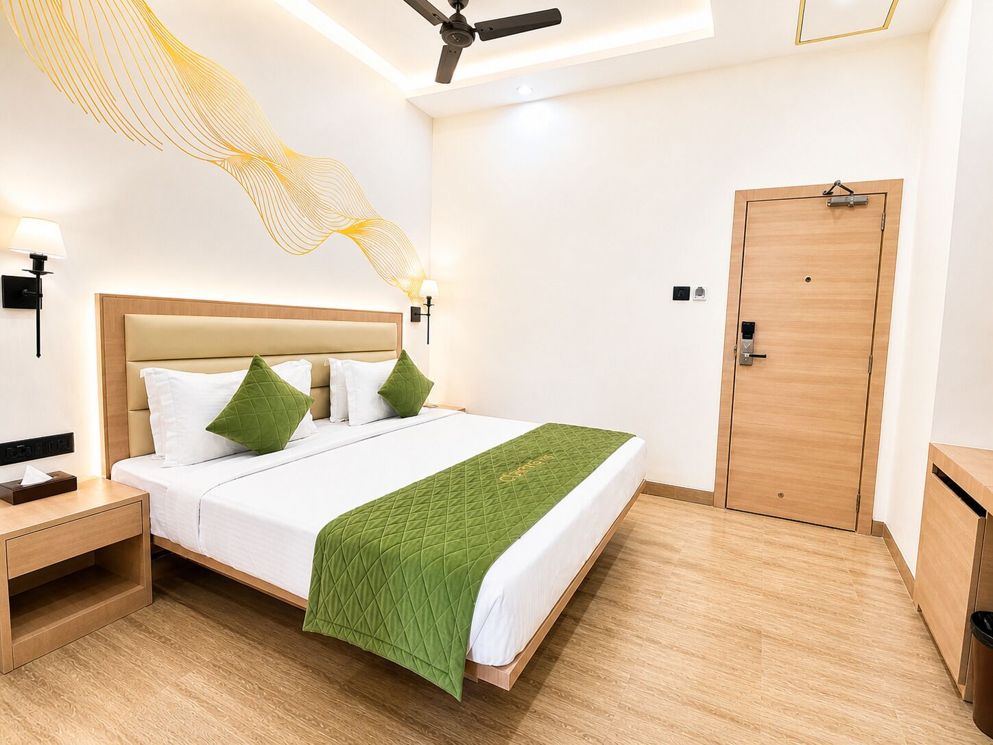 Habitación de hotel con cama, mesita de noche, lámpara de pared, ventilador de techo y puerta de madera.