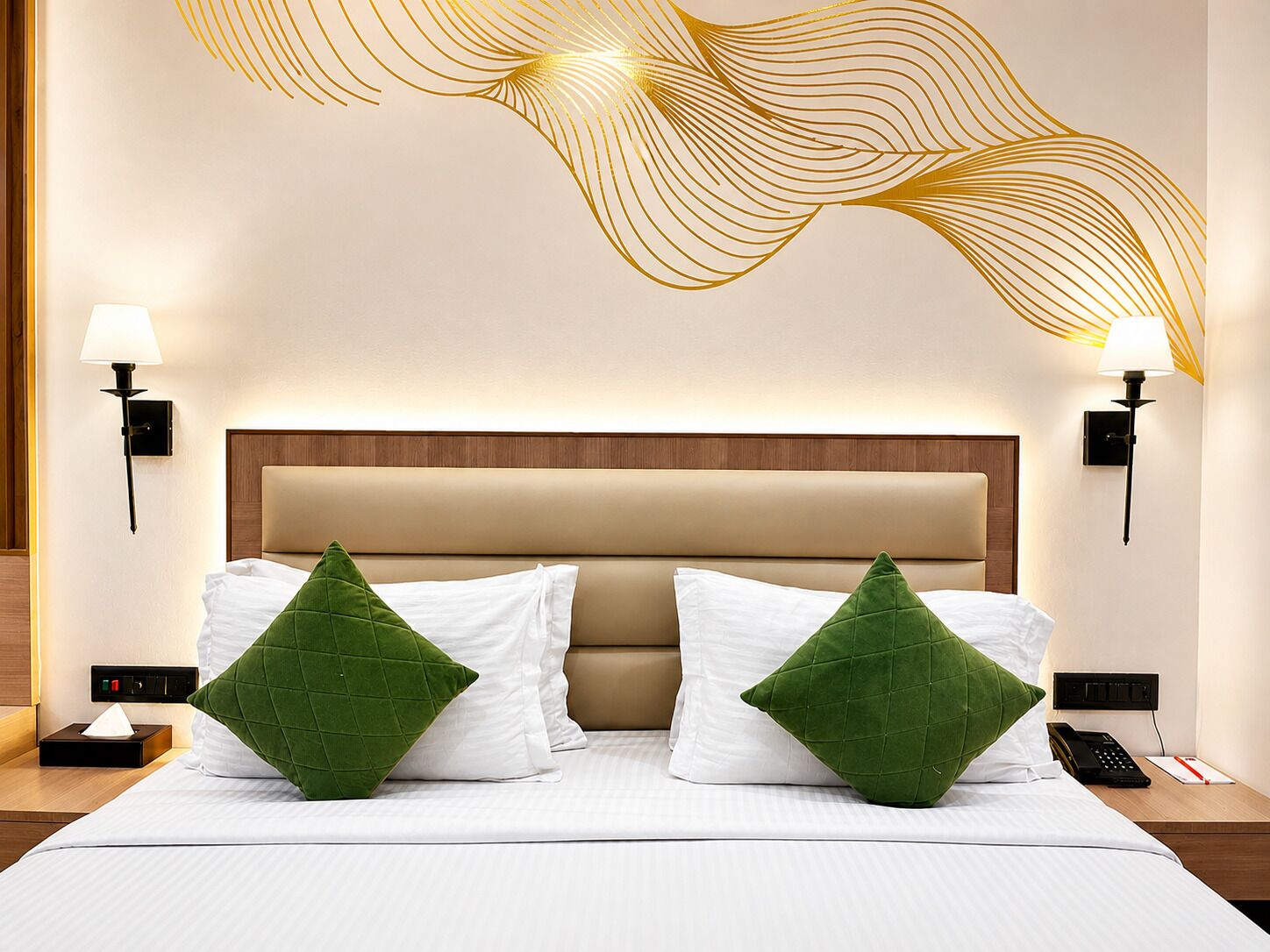A hotel room with a bed, two green pillows, a wooden headboard, bedside lamps, and a wall art piece.