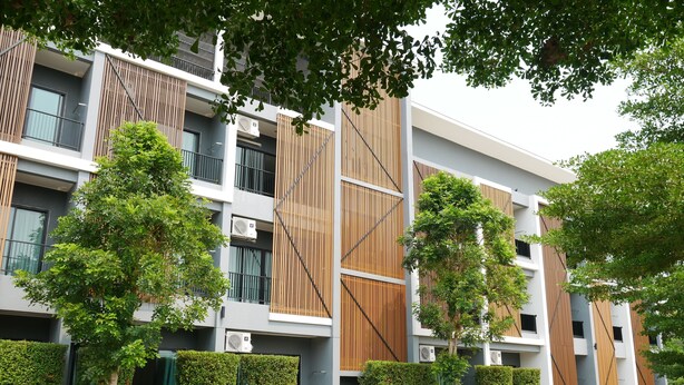 A modern multi-story building with a mix of wooden and metal panel facades, surrounded by well-maintained greenery and a clean, paved ground. - Villa's Day (Phitsanulok)