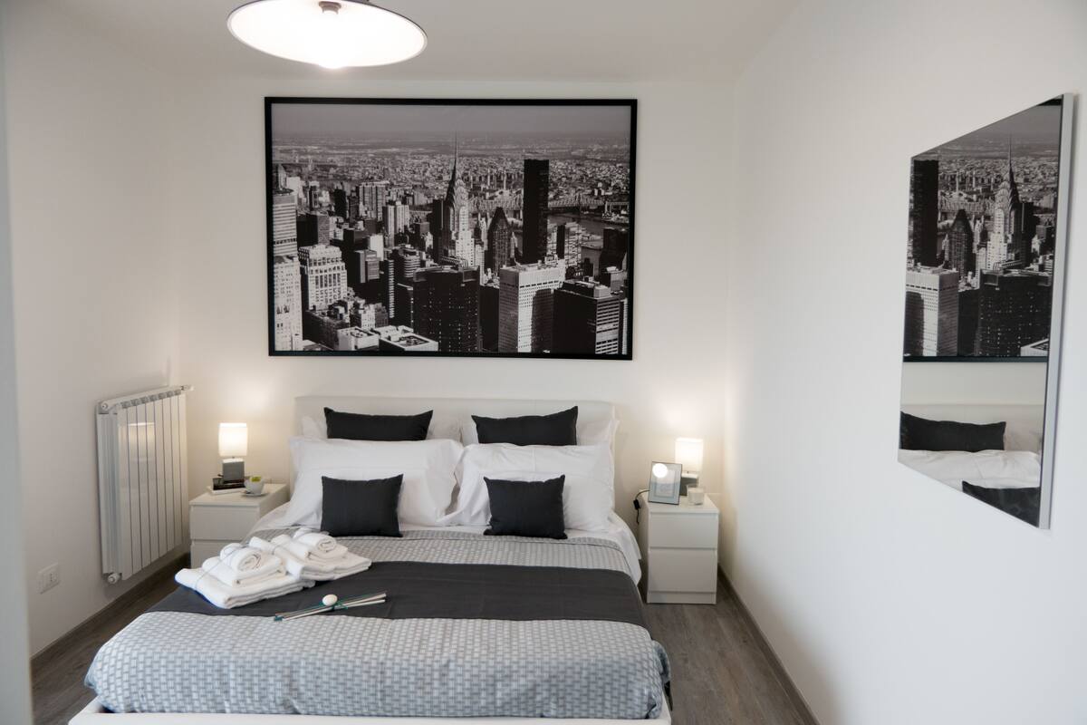 Een moderne hotelkamer met een groot bed, twee nachtkastjes, een radiator en een stadsgezicht kunstwerk boven het bed.