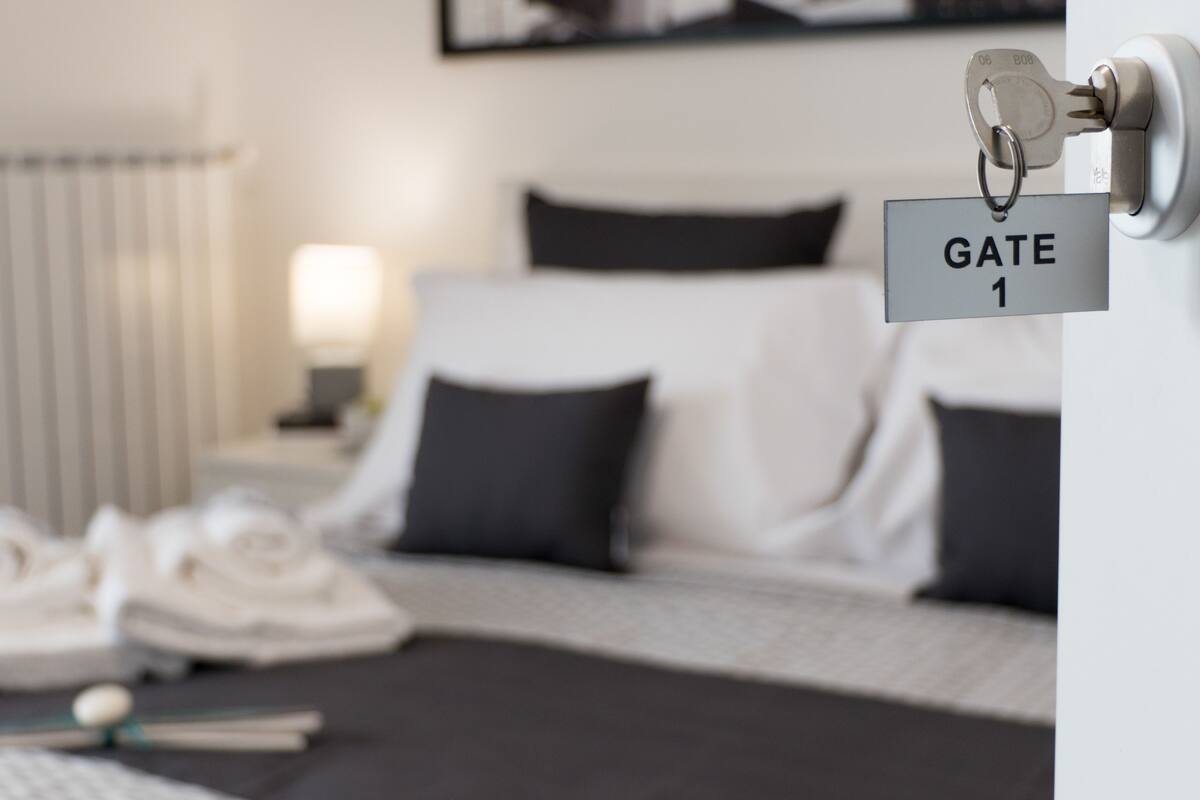 Een hotelkamer met een bed, kussens, een nachtkastje en een sleutelhanger met de tekst 'GATE 1'.