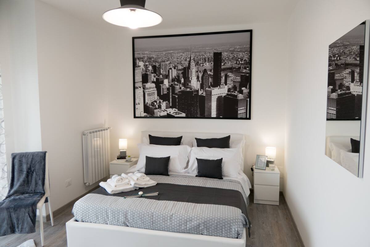 Een moderne hotelkamer met een groot bed, twee nachtkastjes, een stoel, een radiator en een stadsfotografie aan de muur.