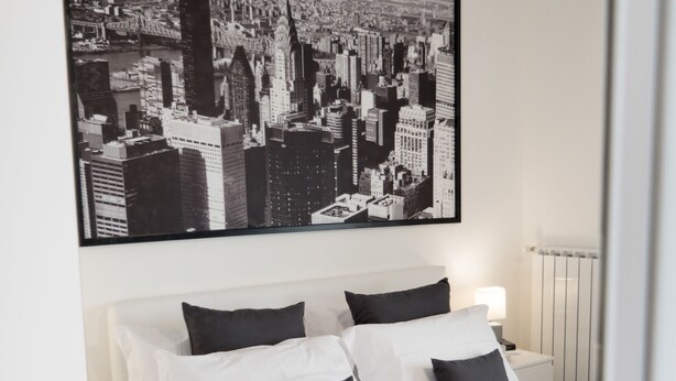 Ein Hotelzimmer mit einem großen Bett, Bettwäsche in Weiß und Schwarz, einem Stadtbild über dem Bett und einem Nachttisch mit einer Lampe.