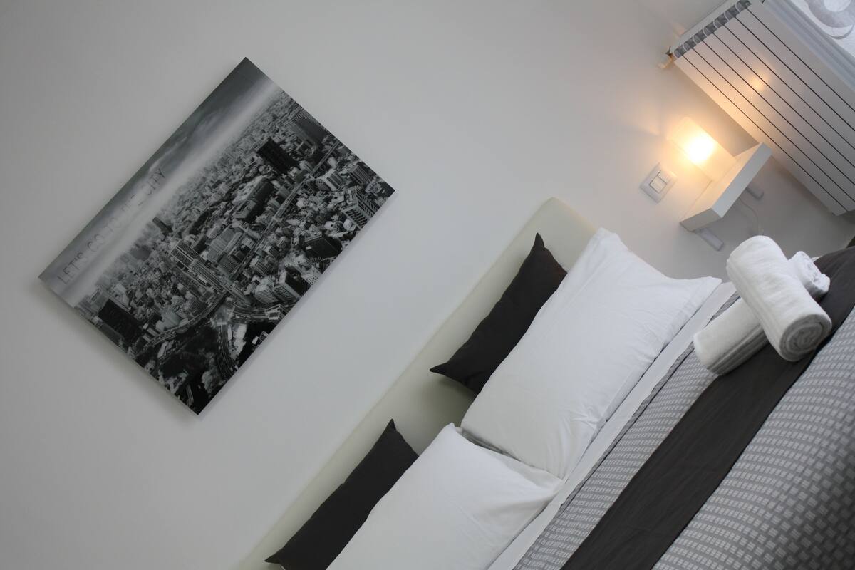 Een opgemaakt bed met witte en donkere kussens, een opgerolde handdoek, en een zwart-wit foto van een stadsgezicht aan de muur.