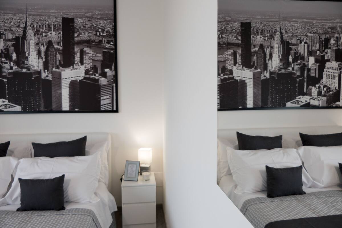 Een hotelkamer met twee bedden, elk voorzien van wit bedlinnen en zwarte kussens. Boven de bedden hangen twee kunstwerken in lijsten, afbeeldingen van een stadsgezicht.