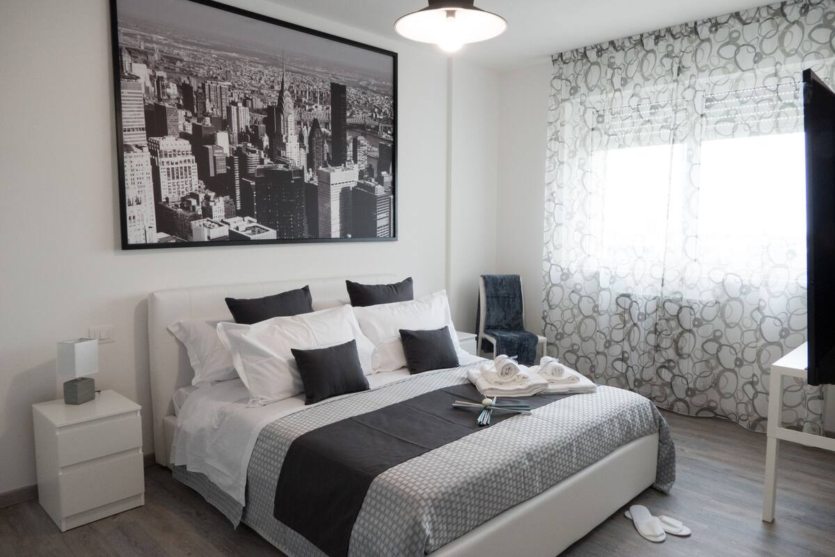 Een moderne slaapkamer met een groot bed, een nachtkastje, een stoel en een stadsgezichtschilderij aan de muur.