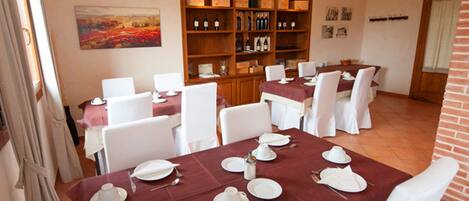 A dining area with tables set for four, white chairs, a wooden wine rack, and a painting on the wall.