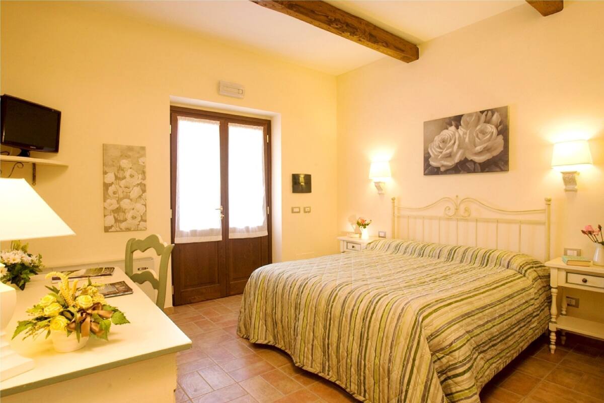 Une chambre à coucher comprenant un lit, des tables de chevet, un bureau avec une lampe, une chaise, un téléviseur fixé au mur et une porte.