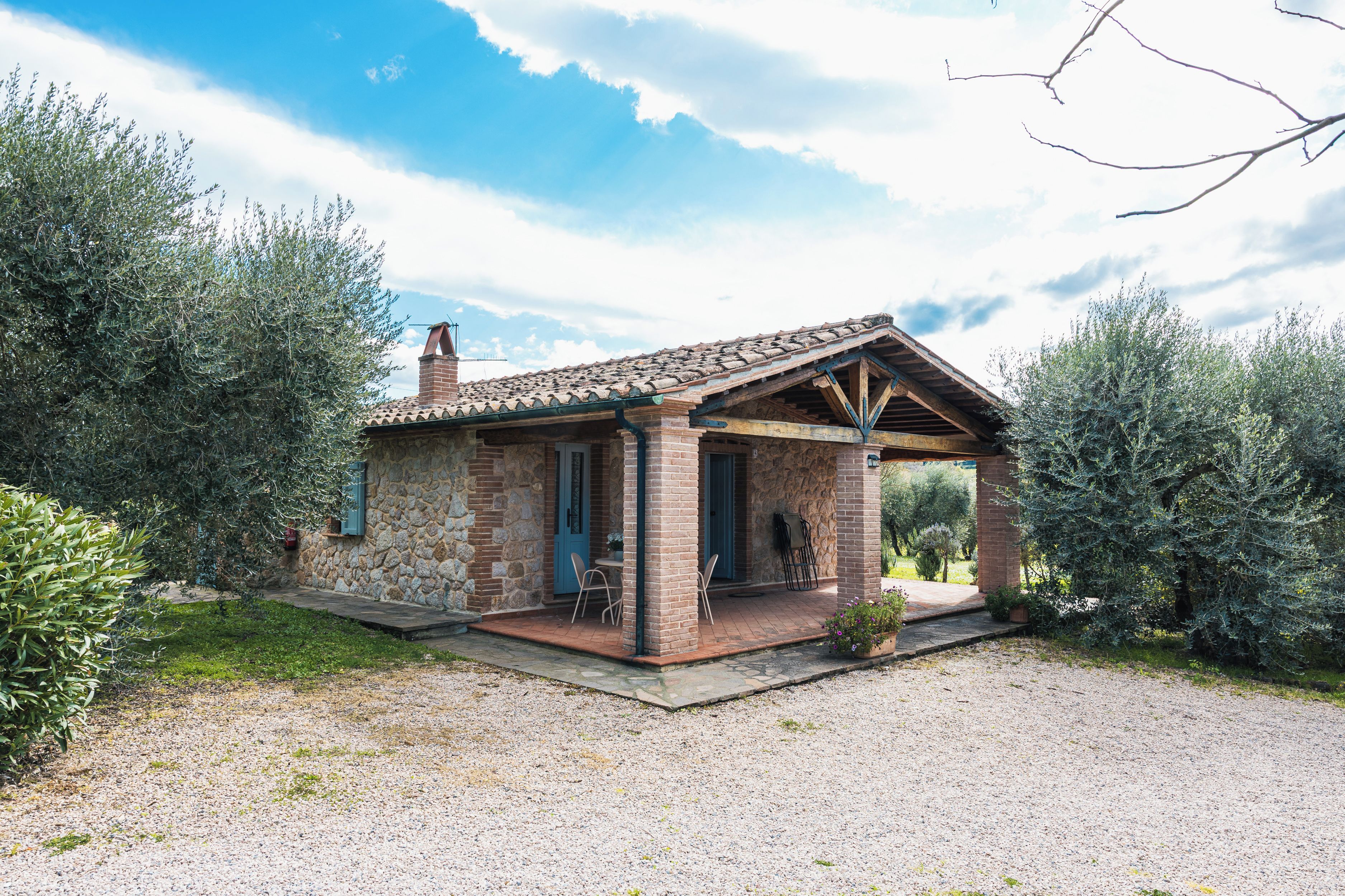 A small stone house with a tiled roof, a porch with chairs, and a gravel driveway.