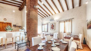 A dining area with a long table set for four, surrounded by white chairs. There is a brick column, a wooden ceiling, and a window with curtains. - Agriturismo Le Cascatelle (Manciano)