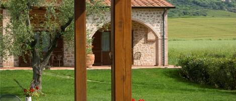 An outdoor patio area with stone flooring, wooden pergola, and a view of a stone building with a tiled roof in the background.