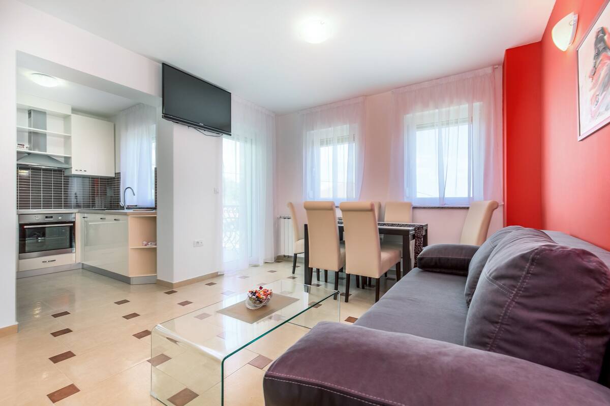 Ein modernes Wohnzimmer mit einem Glastisch, einem violetten Sofa und einem Essbereich mit Stühlen und Tisch.