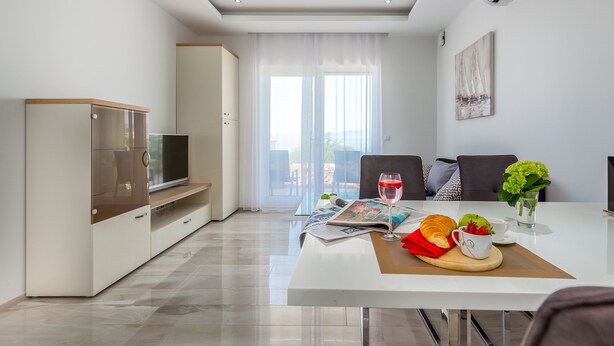 A modern living room with a flat-screen TV, a dining table with a magazine, a glass of red wine, and a croissant. - Luxury Apartments Kostrena (Kostrena)