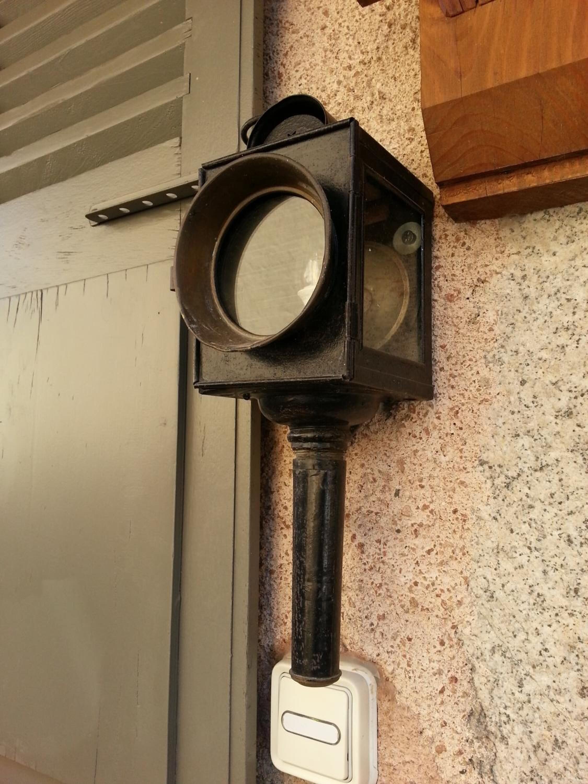 Une lanterne murale dotée d’un miroir rond et d’un interrupteur, fixée à un mur texturé près d’une porte.