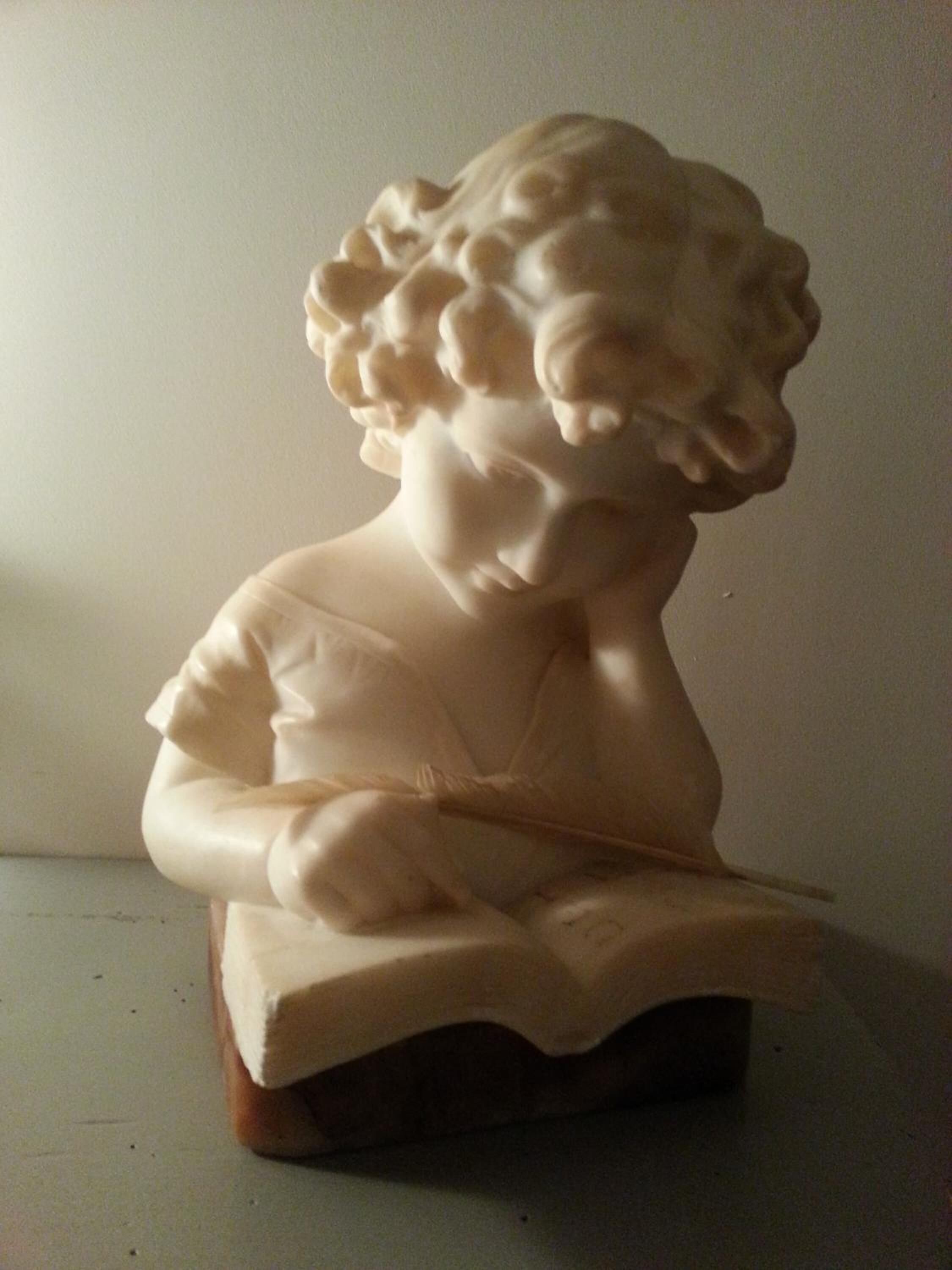 A bust sculpture of a seated figure with curly hair, placed on a wooden base.
