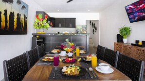 A dining area with a wooden table set for breakfast, black chairs, a ceiling fan, and a TV on the wall. - Villa Maviba Seminyak (Seminyak)