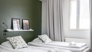 Una habitación con dos camas individuales, un estante blanco con fotografías, una ventana con cortinas y suelo de madera.