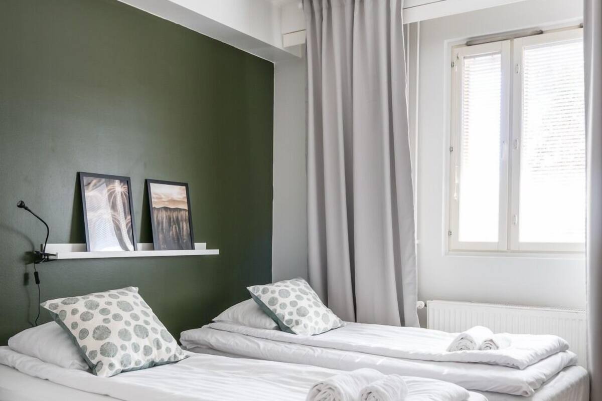 A room with two single beds, a white shelf with pictures, a window with curtains, and a wooden floor.