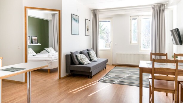 Una sala moderna con un sofá gris, una mesa de comedor de madera y sillas. Hay un televisor montado en la pared y una ventana con cortinas.