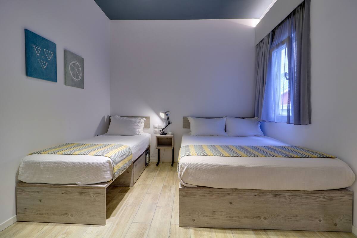 Quarto com duas camas, uma mesa de cabeceira e janela com cortinas.