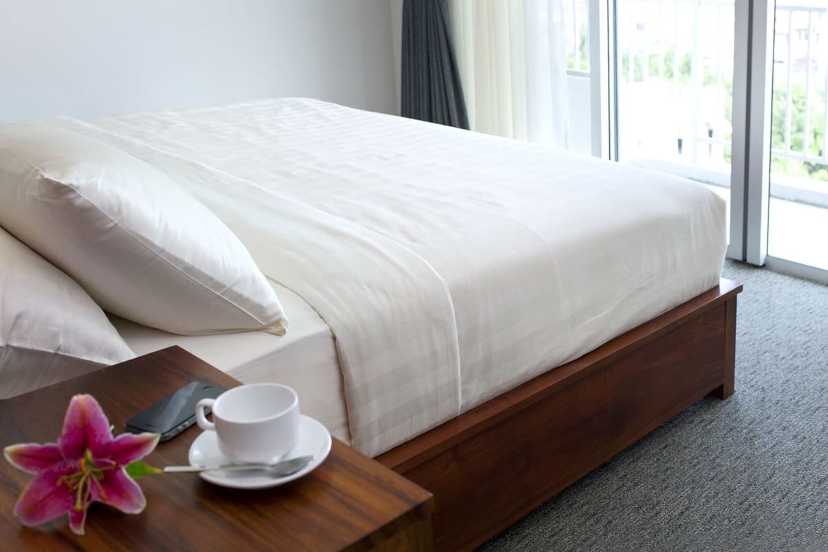 A bed with a wooden frame, a white bedspread, and pillows. There is a wooden nightstand with a white cup and saucer, a spoon, and a purple flower.