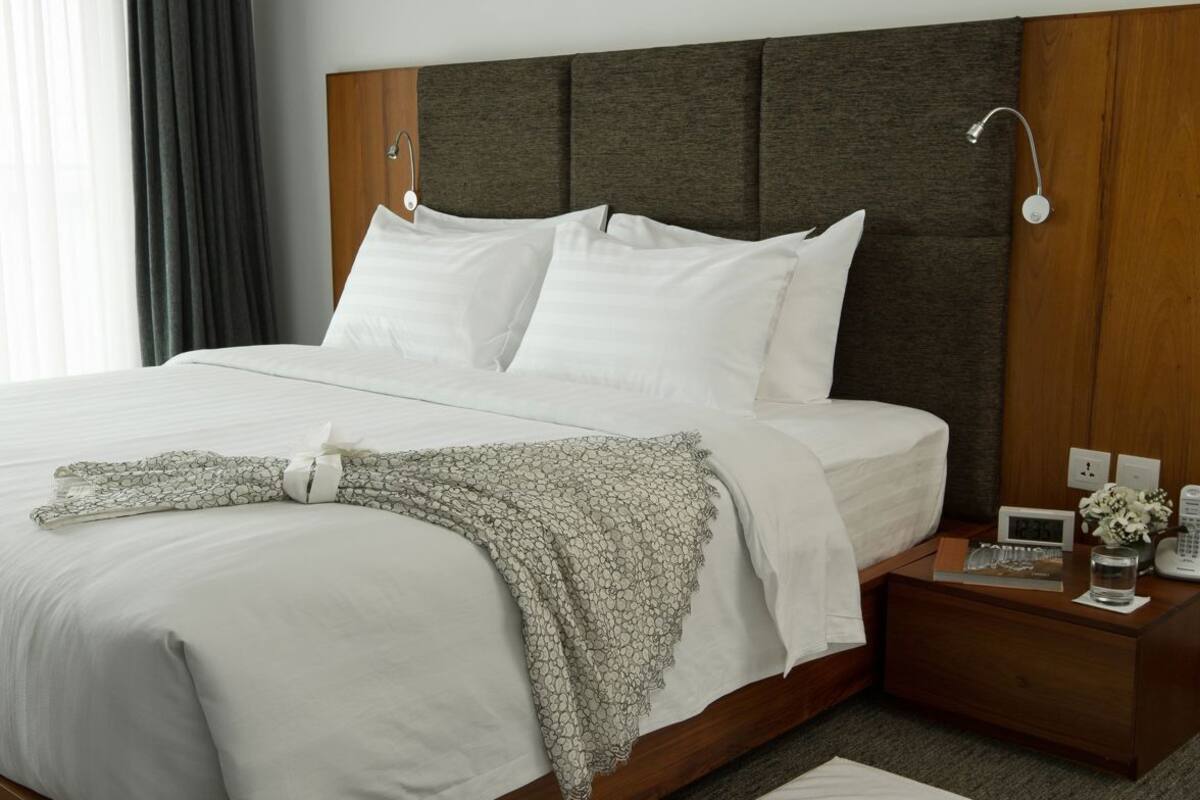A modern hotel room with a large bed, a bedside table with a lamp, a clock, and a vase with flowers.