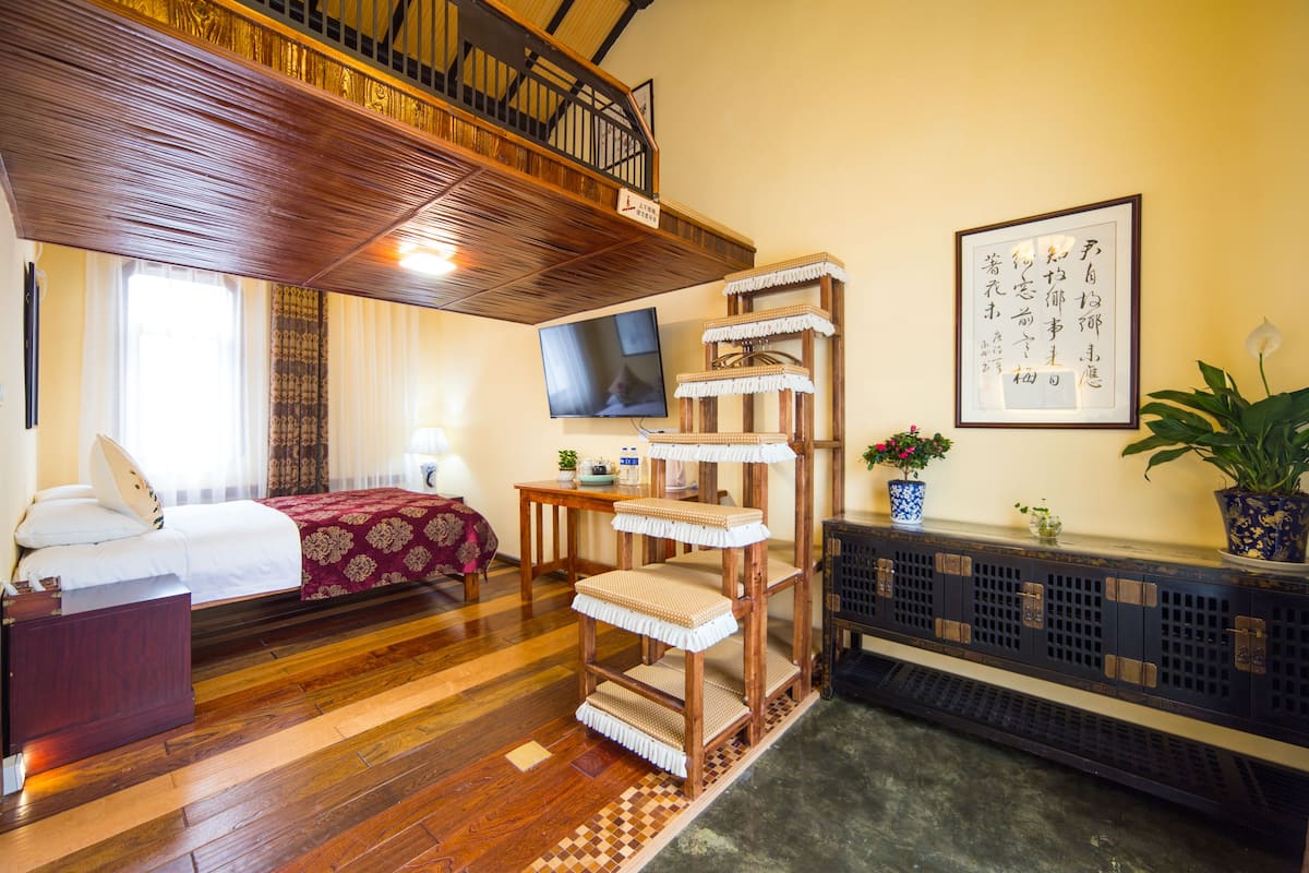 Habitación de hotel con piso de madera, cama con colcha roja y blanca, árbol para gatos de madera, televisión y una obra de caligrafía enmarcada en la pared.
