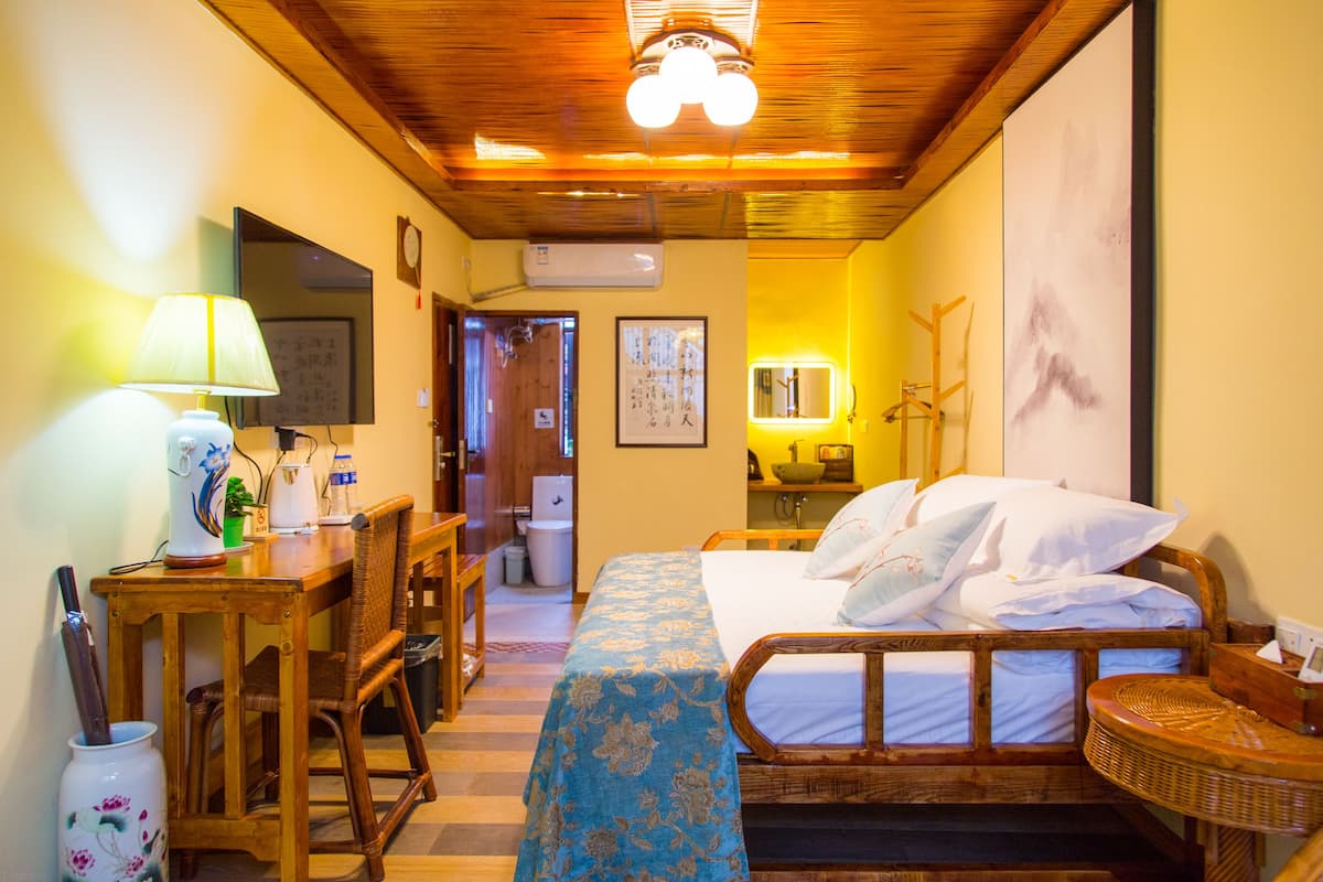 Habitación de hotel con techo de madera, cama con colcha floral azul, escritorio con lámpara, una silla y una pequeña mesa con un florero.