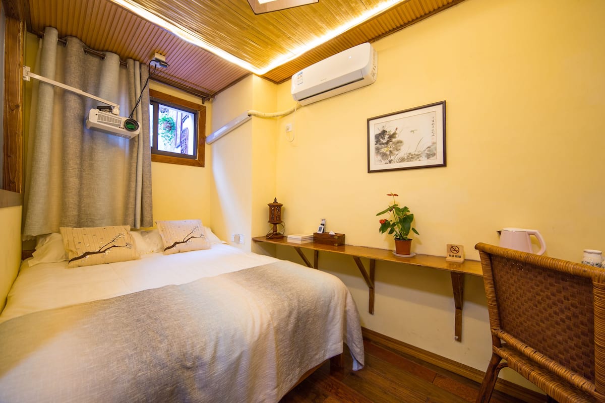 Una habitación de hotel con una cama, un escritorio, una silla, una planta en maceta y un cuadro en la pared.
