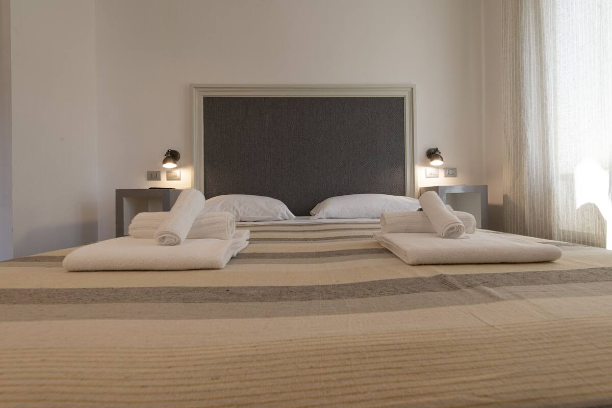 A hotel room with a large bed, two bedside tables with lamps, and a view of the outdoors through sheer curtains.
