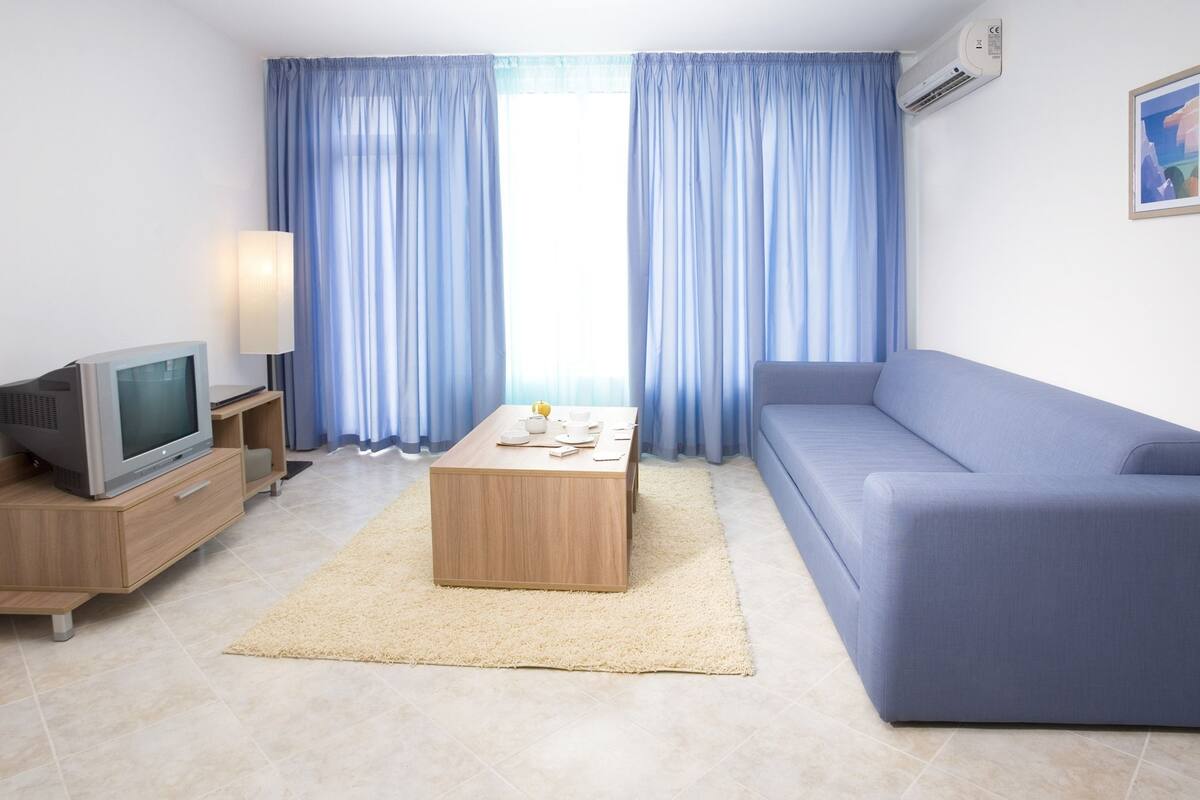 A modern living room with a flat-screen TV, a wooden coffee table, a blue sofa, and light blue curtains.