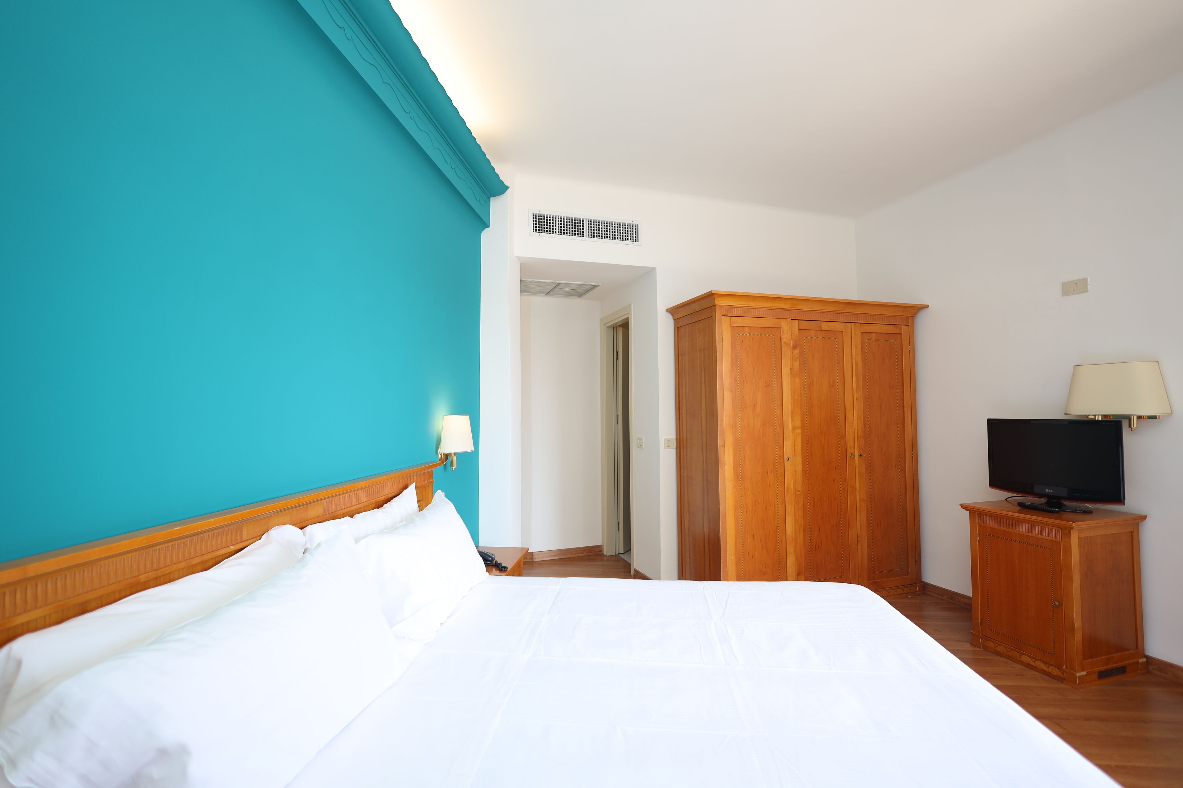 Habitación de hotel con cama, armario de madera, televisor sobre un mueble bajo y puerta que da a otra habitación.