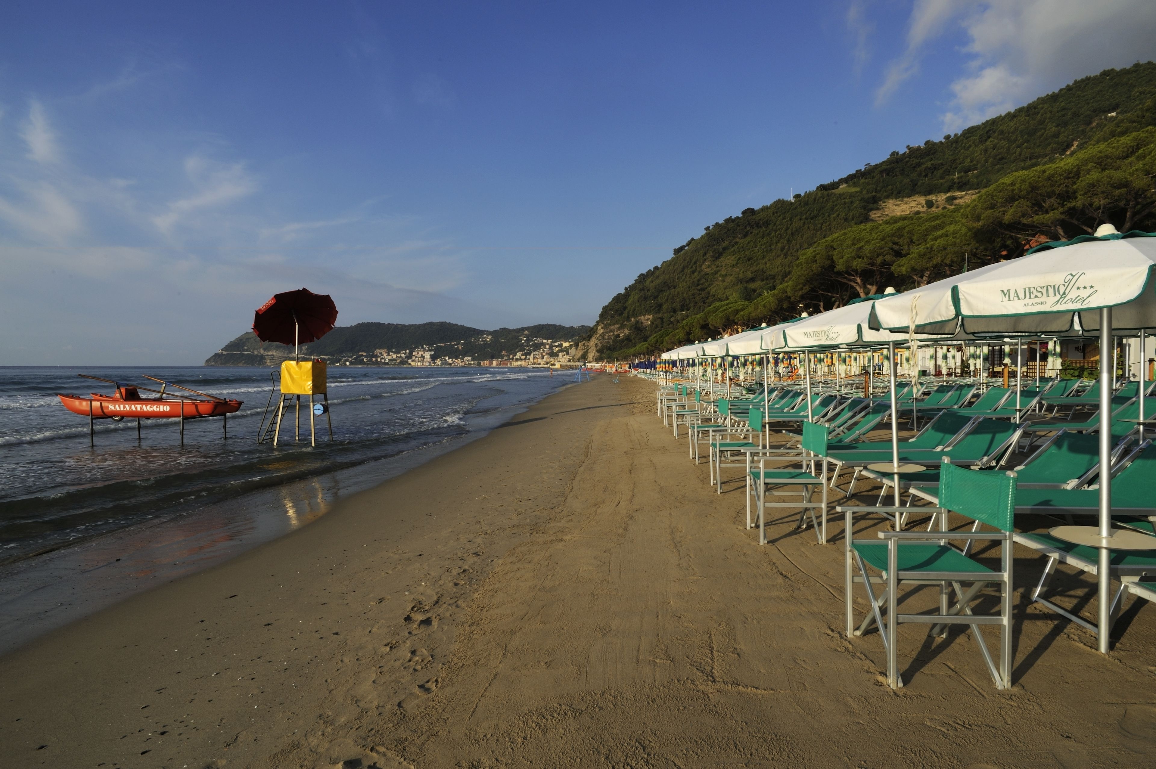 A beach with rows of green lounge chairs under white umbrellas, a lifeguard stand, and a red boat labeled "NAZATAGGIO" on the sand.