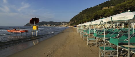 A beach with rows of green lounge chairs under white umbrellas, a lifeguard stand, and a red boat labeled "NAZATAGGIO" on the sand.