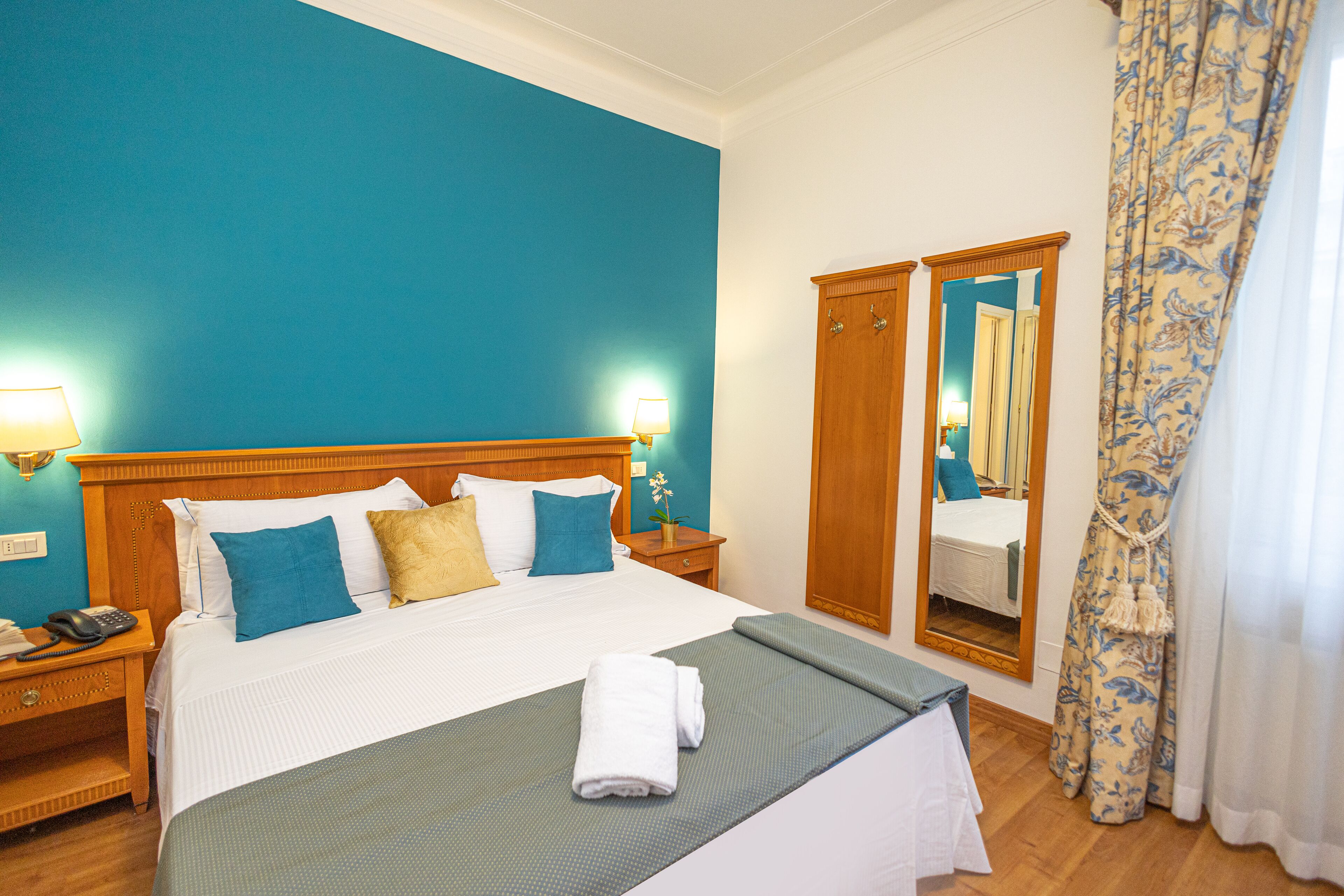 Una habitación de hotel con una cama grande, cabecera de madera, dos mesitas de noche con lámparas, un espejo y una cortina estampada con motivos florales.