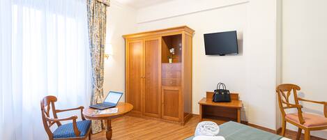 A hotel room with a bed, a desk with a laptop, two chairs, a wardrobe, a television, and a window with curtains.