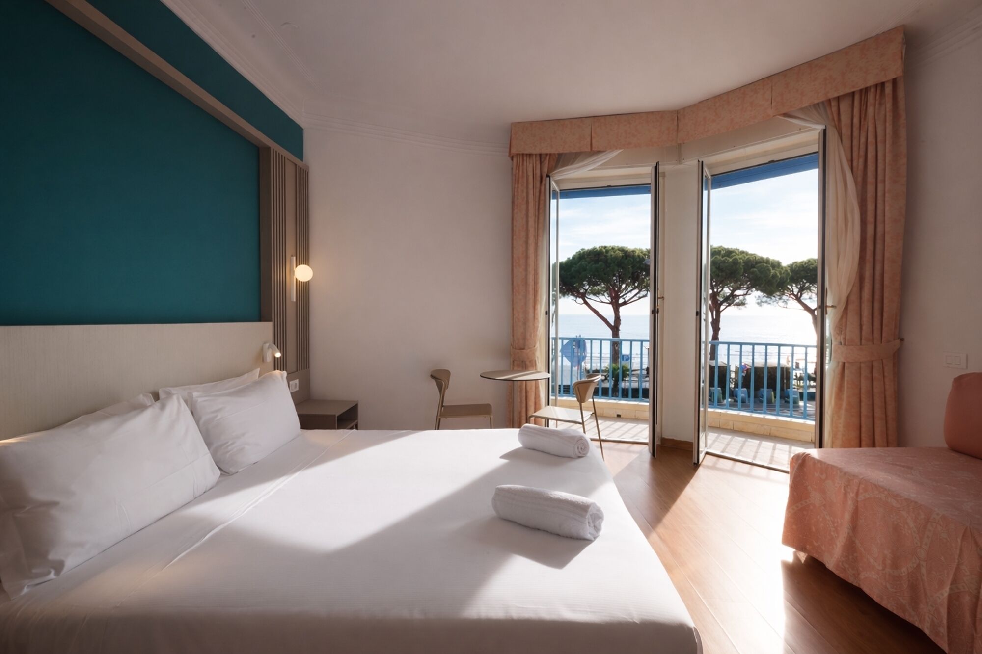 Een hotelkamer met een bed, een kleintje tafeltje, een stoel en een balkon met uitzicht op zee.