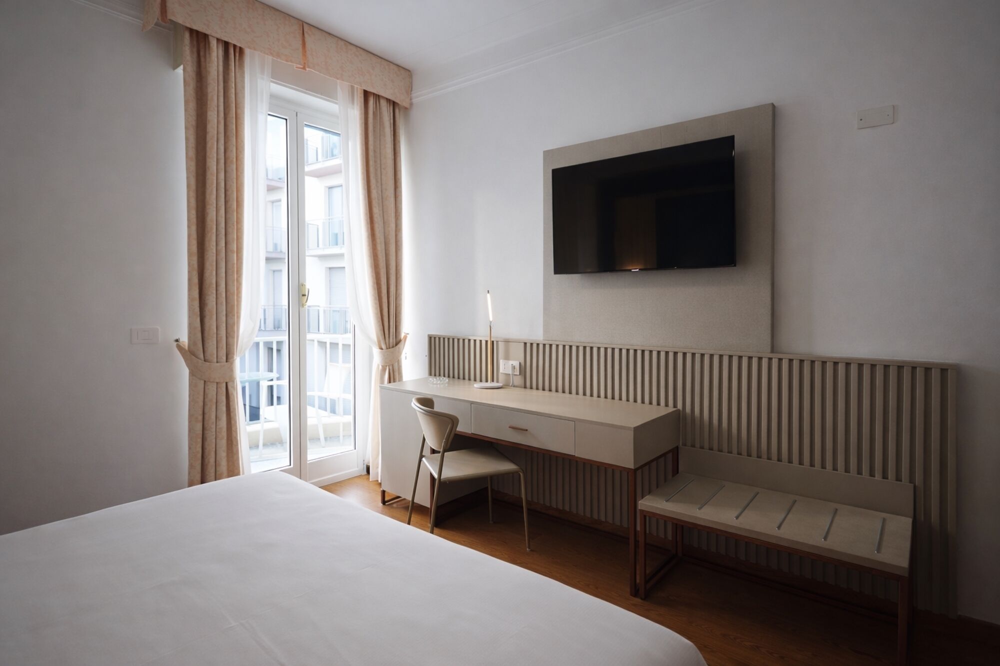 Habitación de hotel con cama, escritorio con silla, televisor montado en la pared y vistas al exterior a través de una ventana.