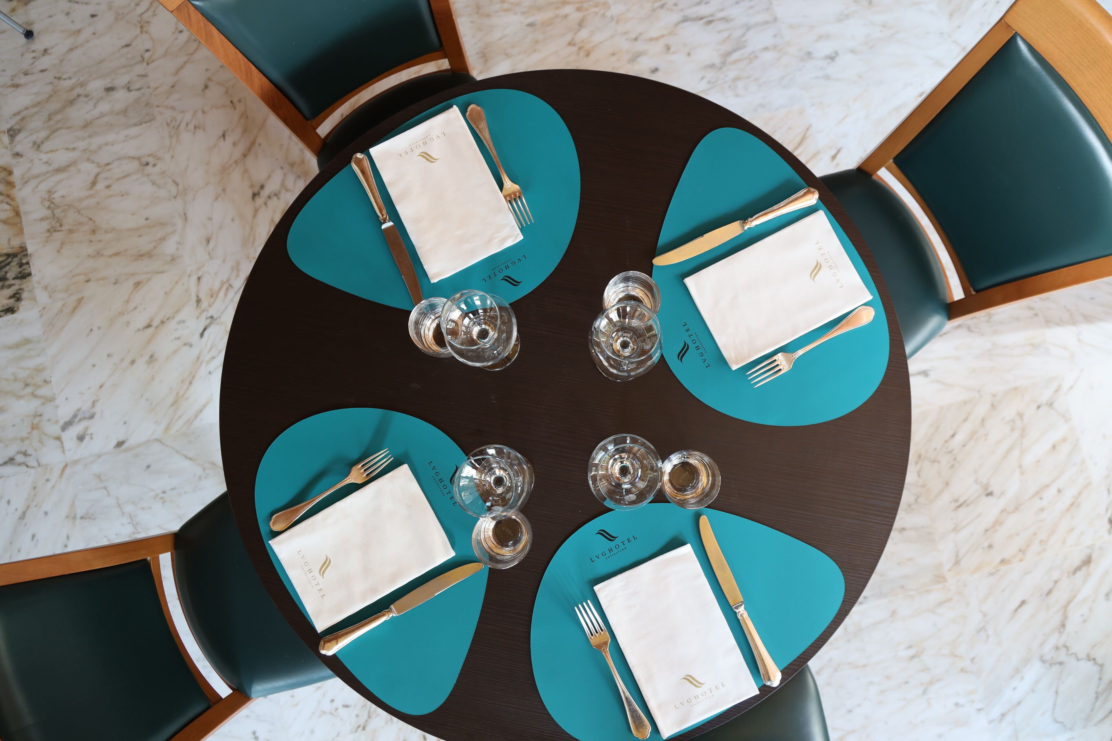 Une table ronde, agrémentée de sets de table et de serviettes blanches de couleur turquoise, avec couverts et verres, et entourée de chaises turquoise.