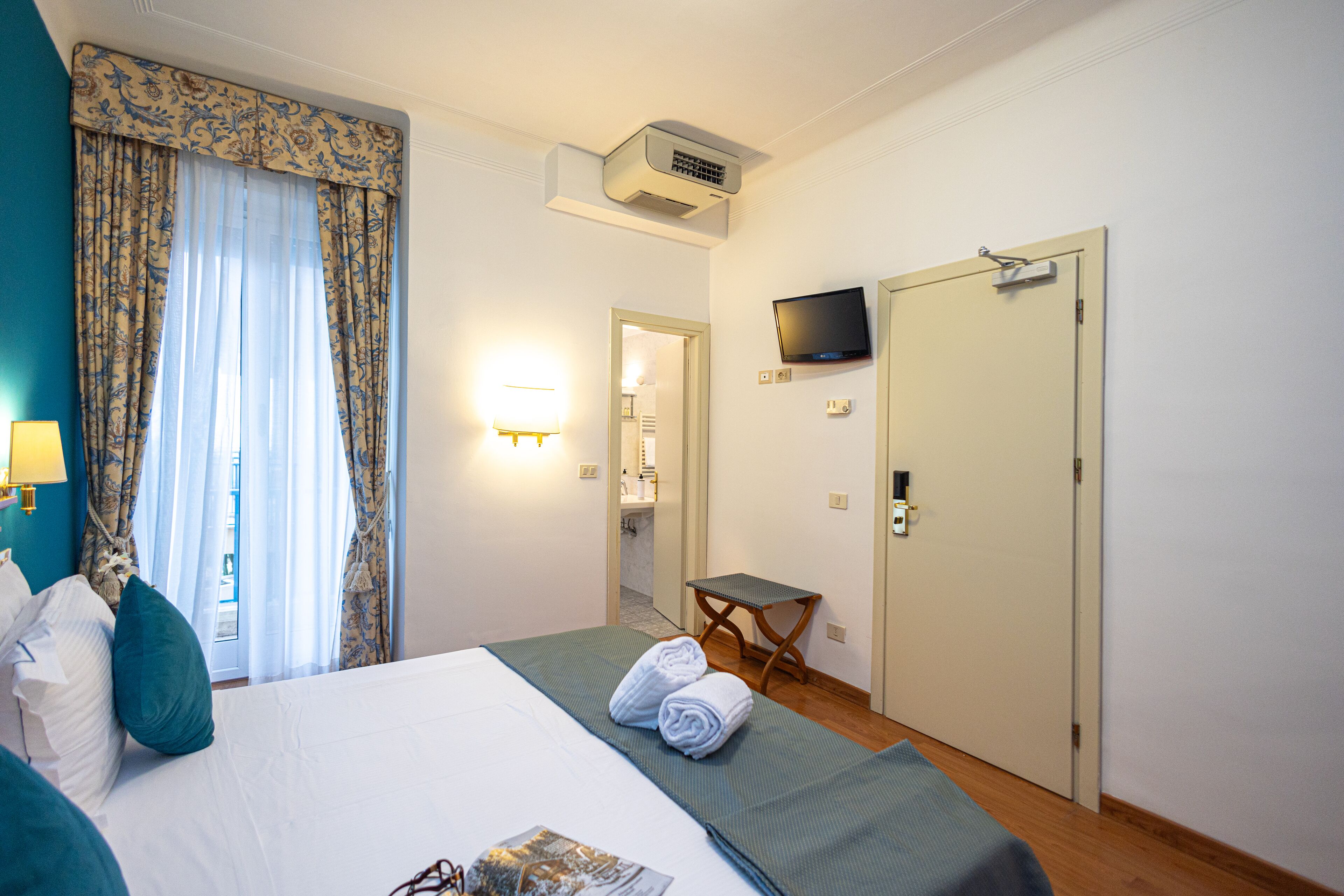 A hotel room with a bed, a TV mounted on the wall, a door, and a window with curtains.