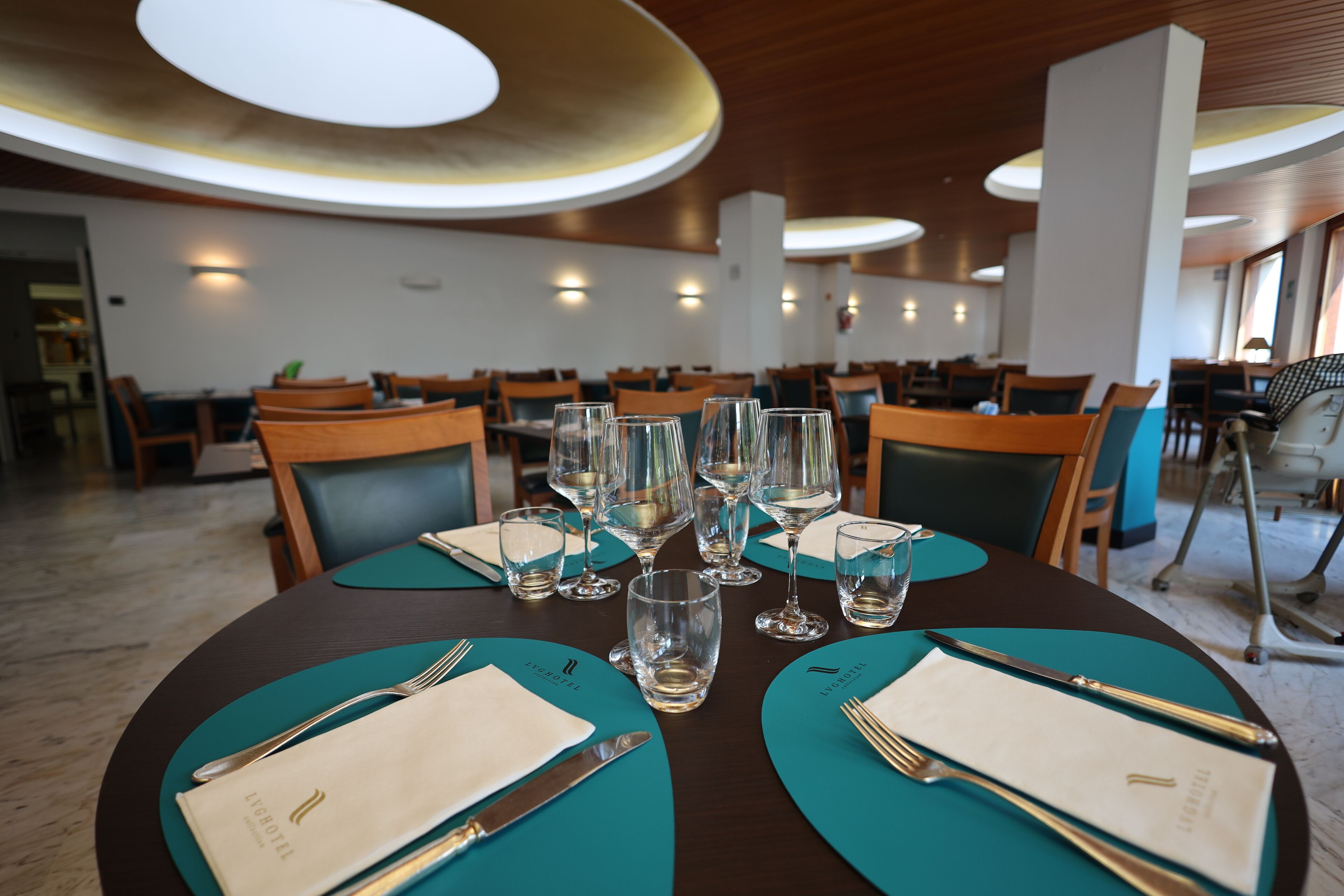 A dining area with tables set for a meal, featuring teal placemats, silverware, and wine glasses.