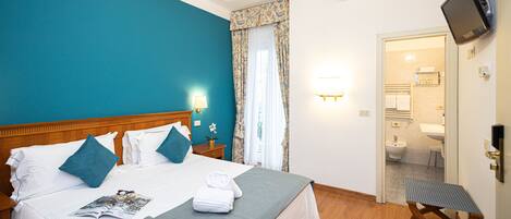 A hotel room with a large bed, a wooden headboard, a bedside table with a lamp, and a bathroom visible through an open door.