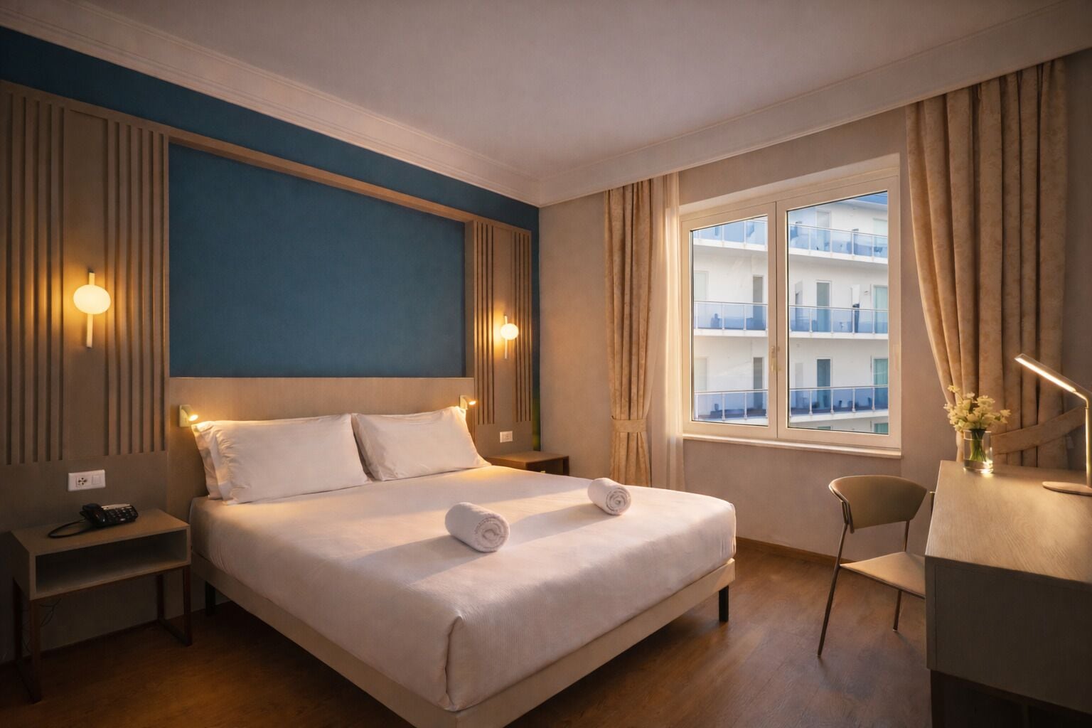 A hotel room with a bed, bedside tables, a desk, and a window with a view of a building.