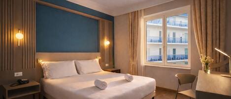Ein Hotelzimmer mit Bett, Nachttischen, einem Schreibtisch und einem Fenster mit Blick auf ein Gebäude.