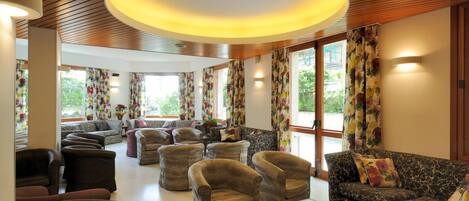 A spacious living area with multiple sofas and armchairs, large windows with floral curtains, and a wooden ceiling.