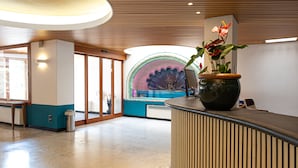 A reception area with a curved reception desk, a potted plant, and a view of a colorful mural in the background. - LVG Hotel Collection - Majestic (Alassio)