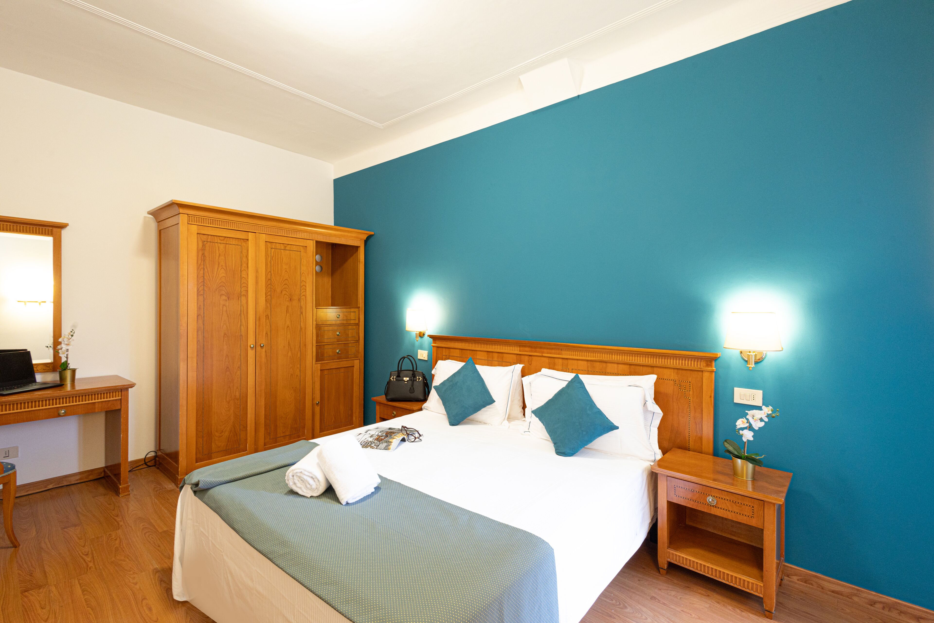 Ein Hotelzimmer mit einem großen Bett, einem Holzschrank, einem Nachttisch und einem Schreibtisch mit einem Laptop.